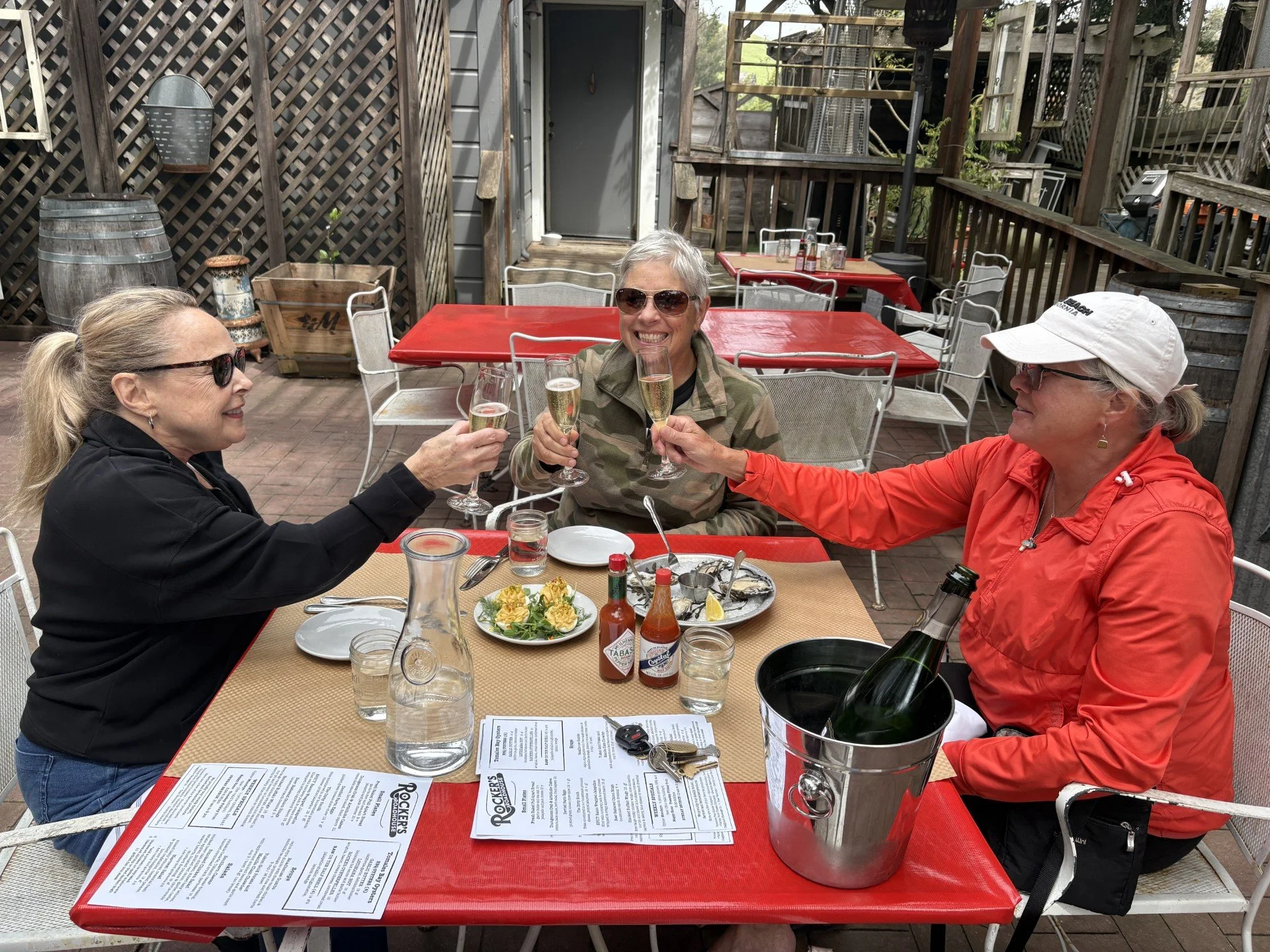 Three women enjoying a meal and drinks at an outdoor restaurant with a red table, surrounded by wooden fencing and patio furniture.