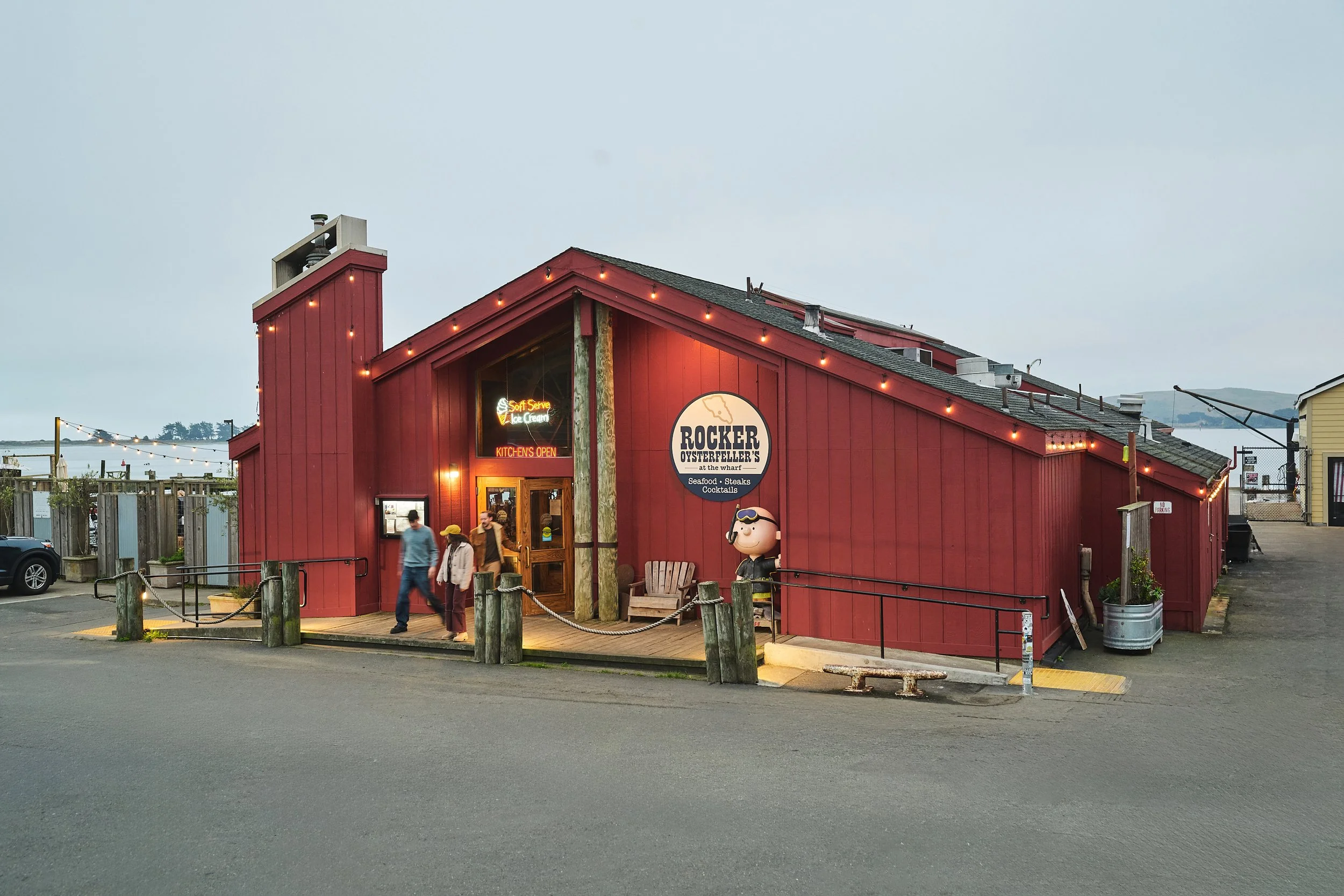 Red seafood restaurant called Rocker Oysterfeller's with string lights, neon sign, and outdoor seating area, near water in the background, during evening.