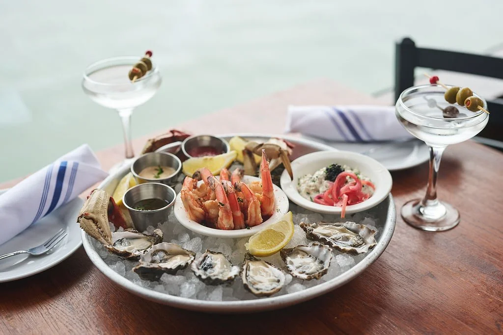Seafood platter with oysters on ice, shrimp, and dipping sauces, garnished with lemon wedges, served with two cocktail drinks garnished with olives and cherry tomatoes.