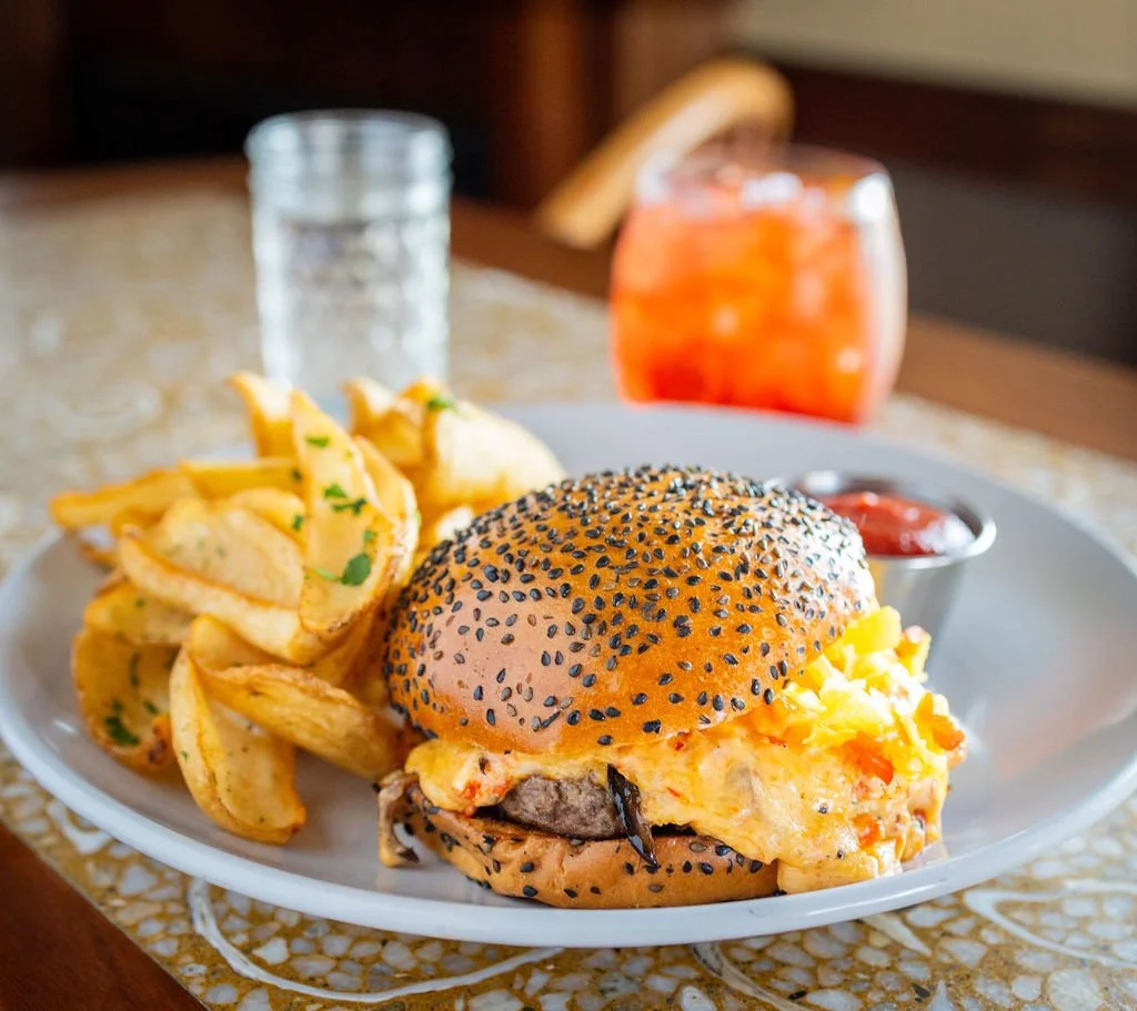 A plate with a cheeseburger sandwich topped with sesame seeds, potato wedges garnished with parsley, ketchup in a small cup, and a glass of iced tea with lemon on the side.