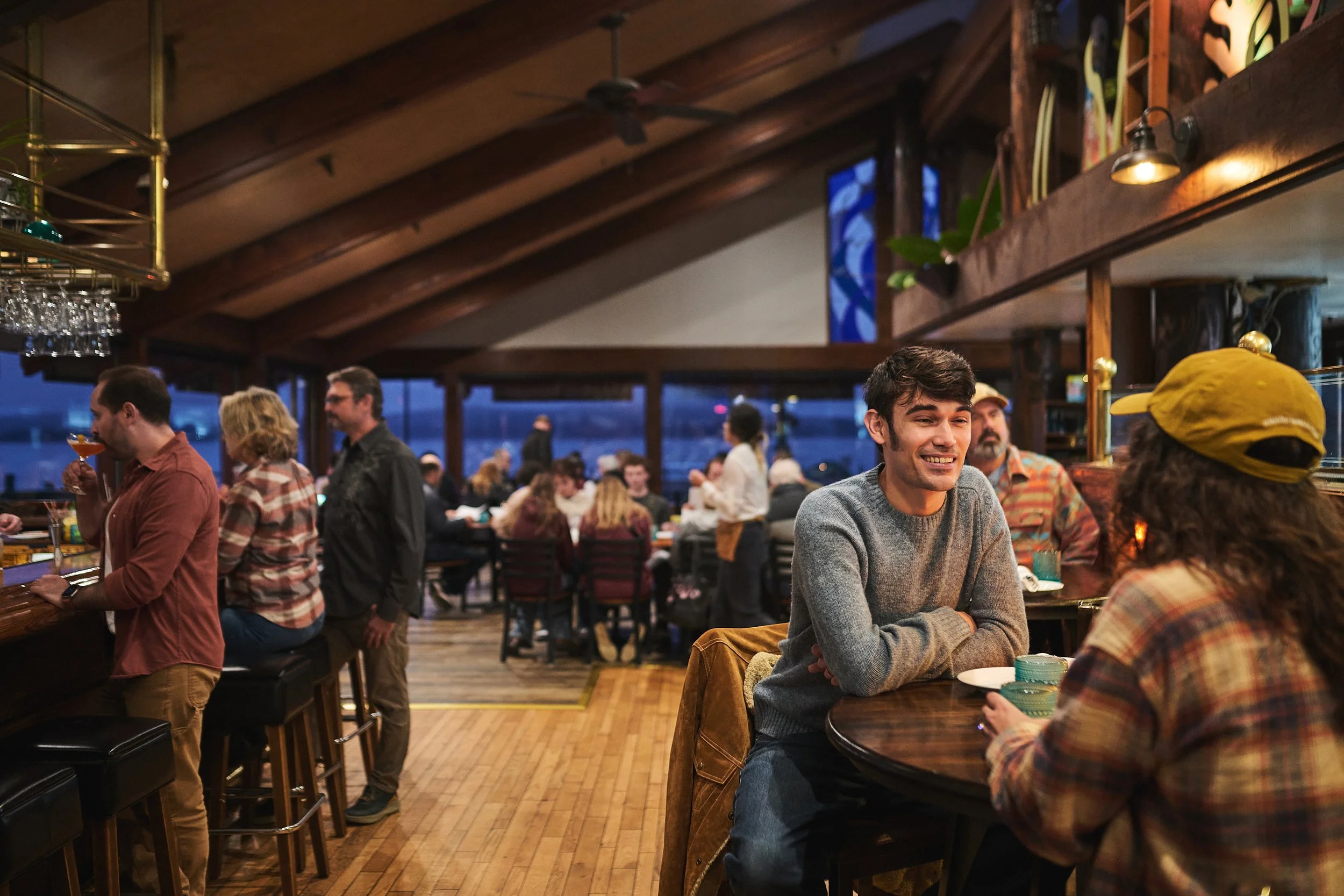 People socializing inside a rustic-style bar or restaurant with wooden beams, large windows, and a bar area, during evening hours.