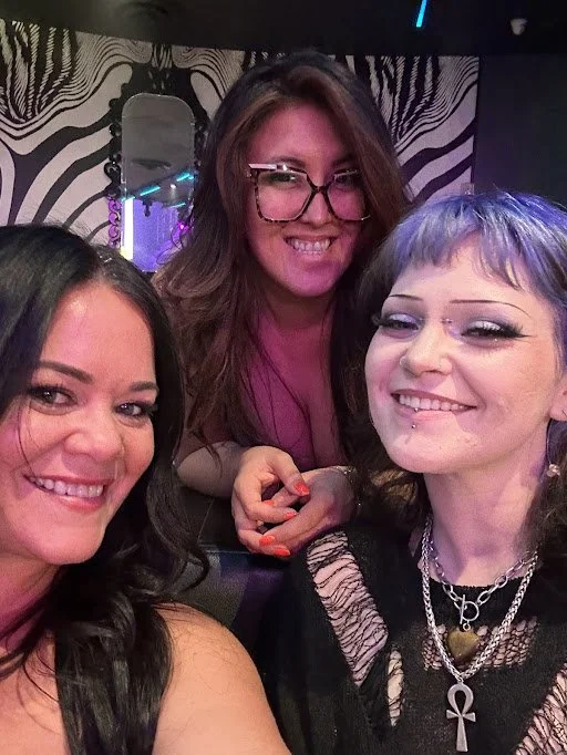 Three women smiling and posing for a selfie in a dimly lit, decorated indoor setting with black and white striped wallpaper and neon lights.