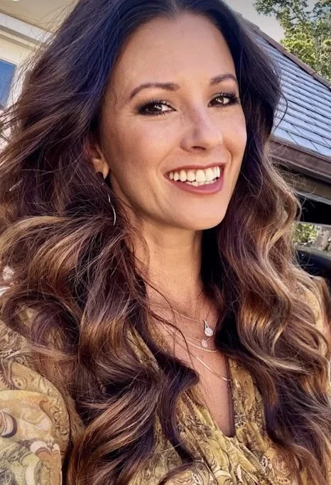 A woman with long, wavy brown hair smiling outdoors with a house and trees in the background.