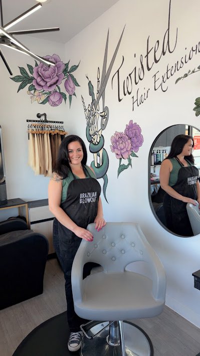 Woman with dark hair and a black apron standing in a hair salon, smiling, with a large mirror behind her. The salon has decorative purple flowers and a wall sign that says 'Twisted Hair Extension'.