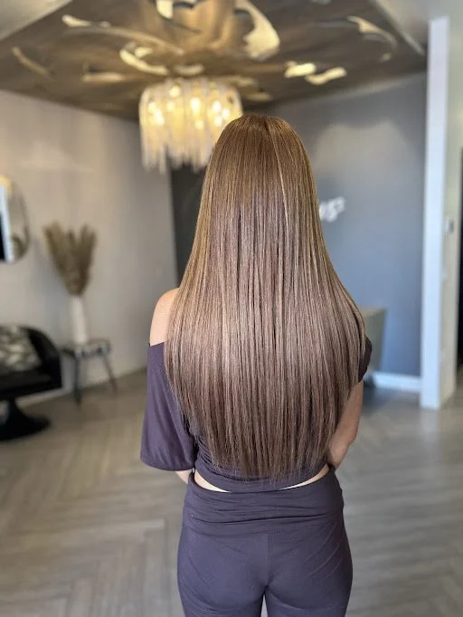 A woman with long, straight, light brown hair standing with her back to the camera in a modern living room.