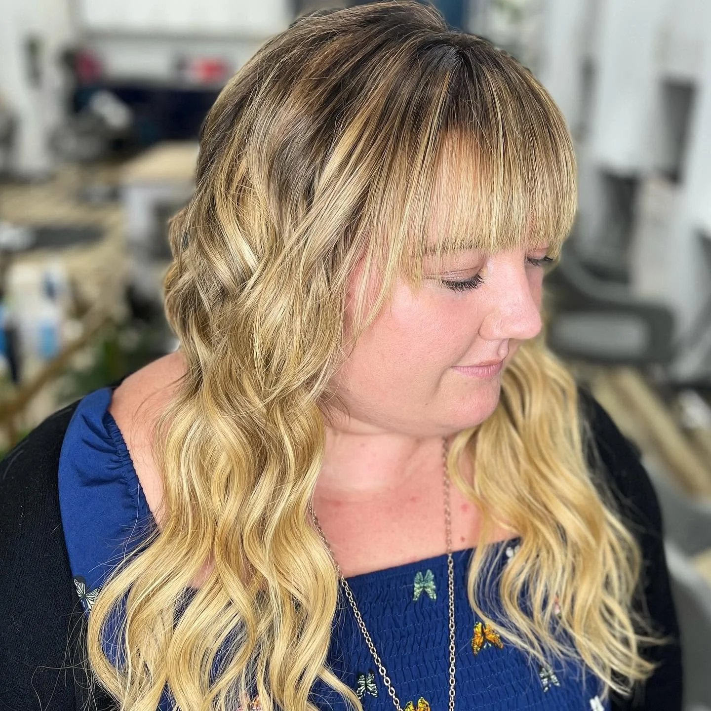A woman with long blonde, wavy hair and blunt bangs, wearing a blue top with butterfly and flower patterns, is looking down with a soft expression in a hair salon.