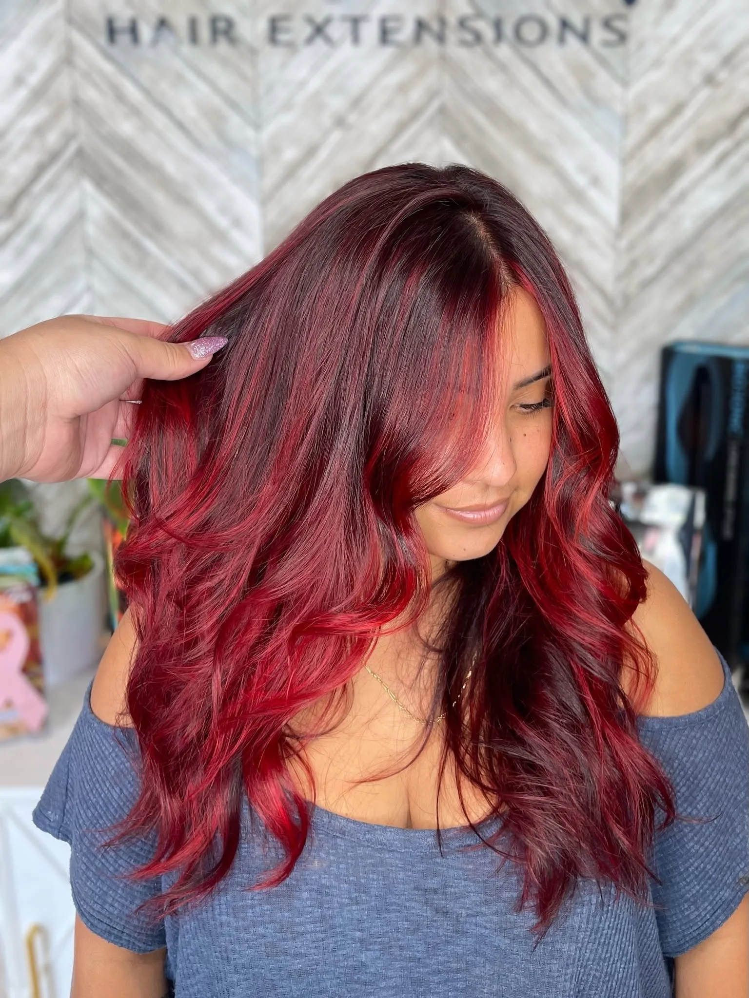 A woman with long, wavy hair dyed in shades of red and burgundy, wearing a blue off-shoulder top, smiling with her eyes closed.