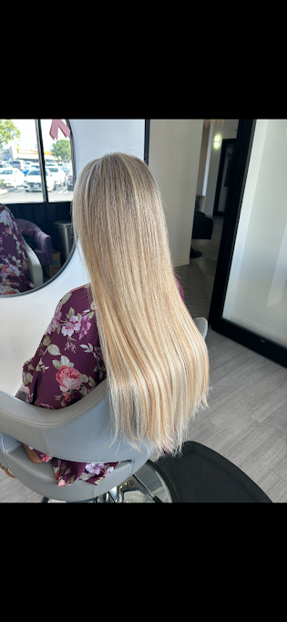 Back of a woman with long, straight, blonde hair sitting in a salon chair wearing a purple floral robe, with a mirror reflecting her and the salon interior.
