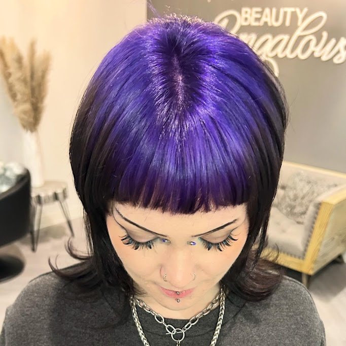 Woman with short black hair and purple highlights, looking down, in a salon with a sign that says 'Beauty' in the background.