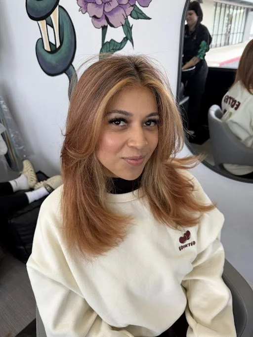 A woman with light brown hair and makeup seated in a salon, with a colorful floral mural behind her and people visible in the background.