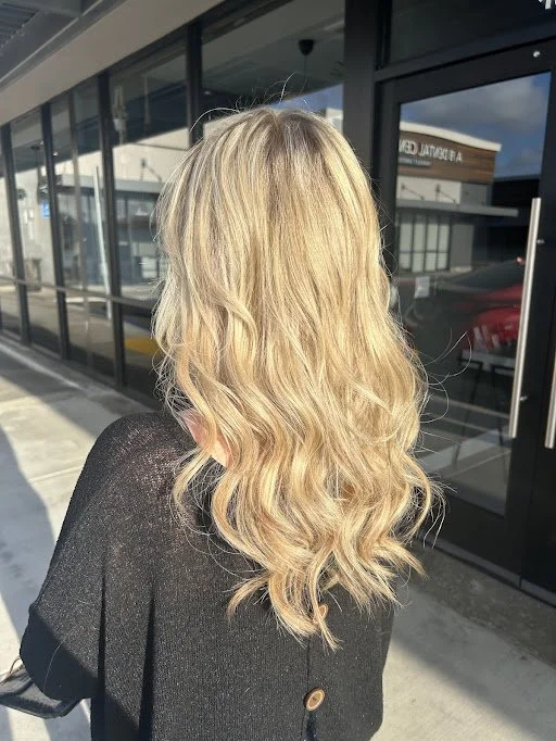 Back of a woman with long, wavy blonde hair standing outside a building with glass windows and a glass door.