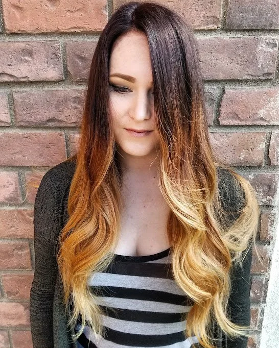 A woman with long, curly hair that is dark at the roots and fades to blonde at the tips, standing against a brick wall.