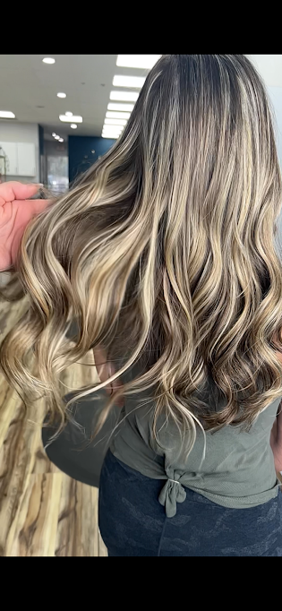 A woman with long, wavy blonde hair showing darker roots, standing in a salon. The background includes salon lighting and mirrors.