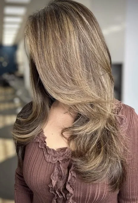 A woman with long, wavy, light brown hair turned to the side, wearing a mauve blouse with ruffles.