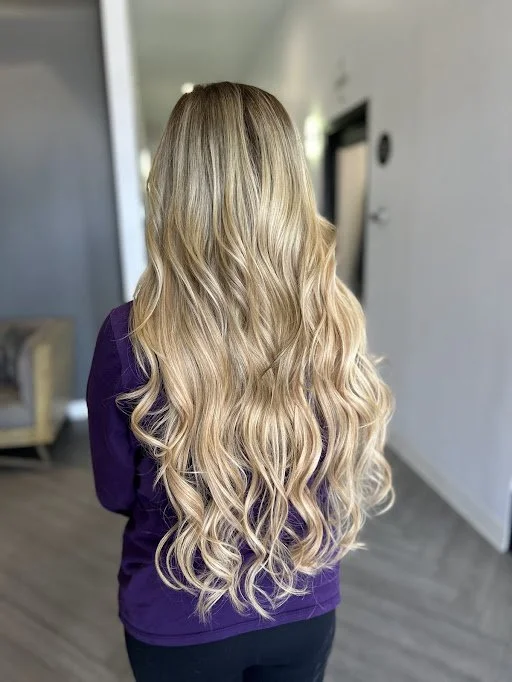 Back view of a woman with long, blonde, wavy hair wearing a purple top in an indoor setting.