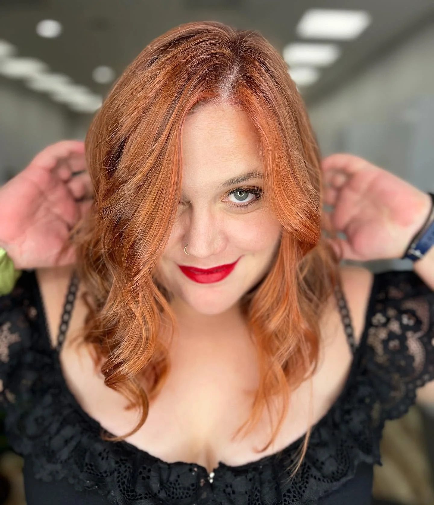 A woman with long, wavy red hair and bright red lipstick, wearing a black lace top, smiling and adjusting her hair in a salon or similar setting.