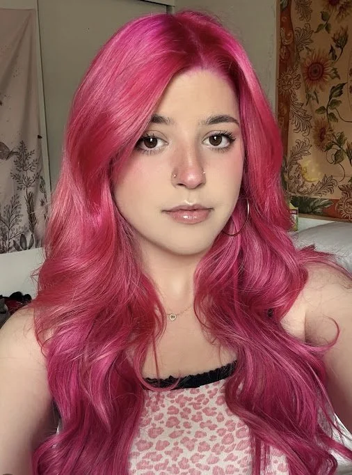 Young woman with long, wavy pink hair, wearing a light pink and black sleeveless top, gold hoop earrings, a small heart necklace, and minimal makeup, in a room with floral curtains and floral wallpaper in the background.