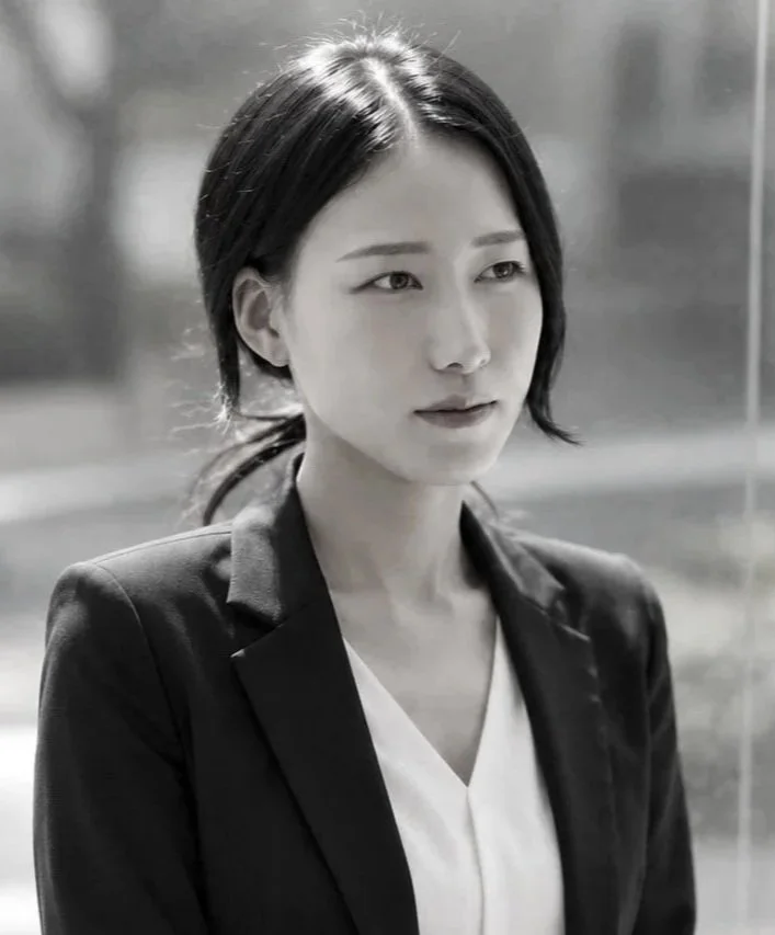 A woman with black hair styled in a ponytail wearing a black blazer and white top, standing outdoors with a blurred background.