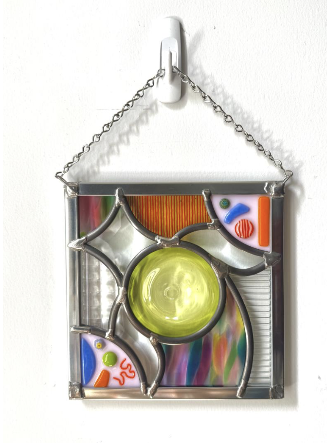 Sharon Whelton Glass Artist - Colorful stained glass wall hanging with abstract designs, small glass pieces, and a yellow central circle, hanging from a white hook on a white wall.