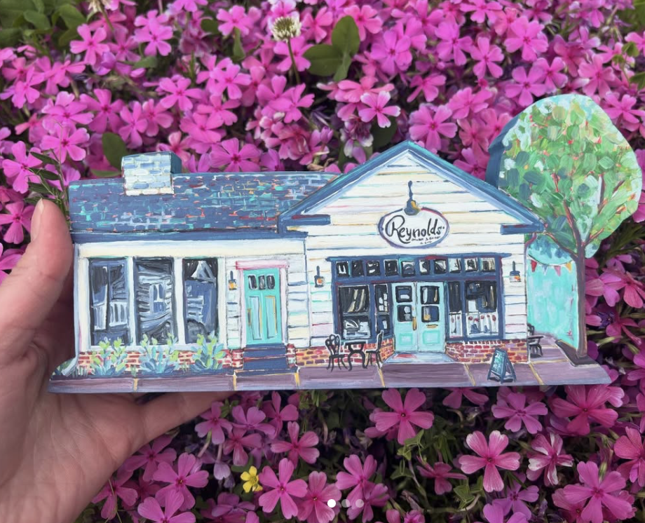 Magpie Maketh - A colorful illustrated model of a storefront named Reynolds, with a porch and outdoor seating, set against a backdrop of pink flowers and a painted tree.