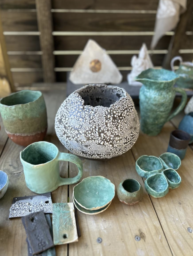 Healing Arts ATL - Various handmade pottery such as mugs, bowls, and vases in shades of green, blue, and earth tones displayed on a wooden table with a rustic wooden background.
