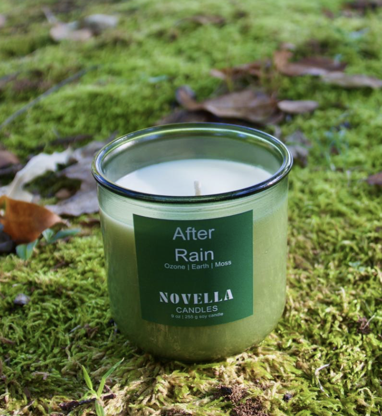 Novella Candles - Green candle in glass jar labeled "After Rain" with scent notes of ozone, earth, and moss, placed on mossy ground with scattered leaves in an outdoor setting.