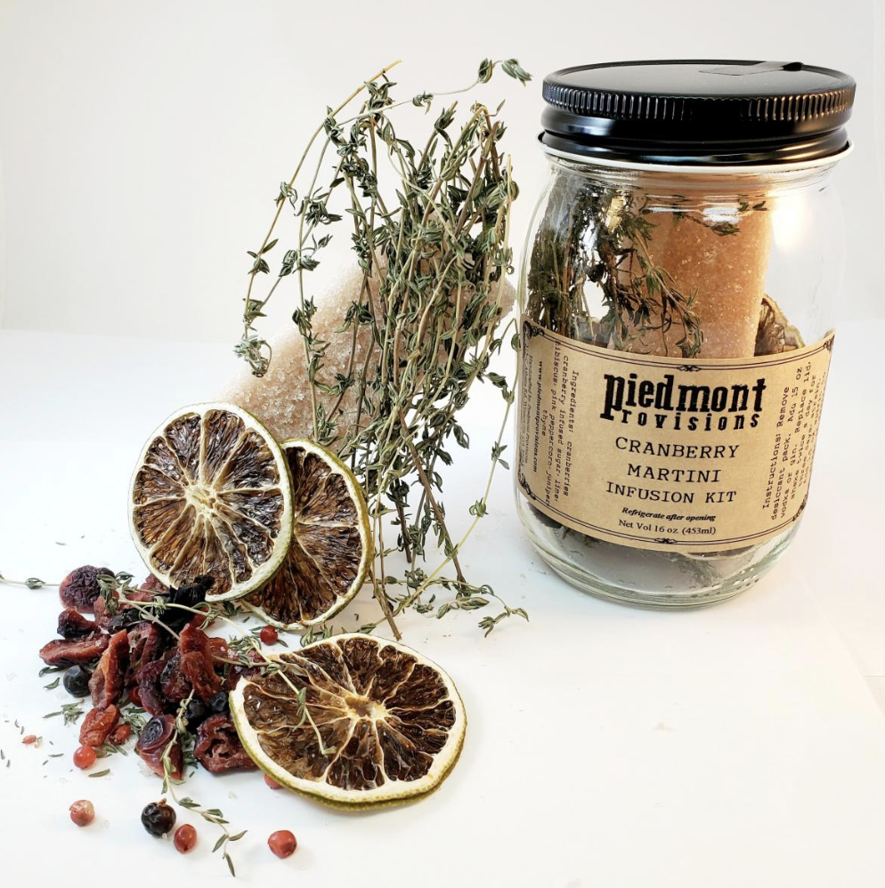 A jar labeled 'Piedmont Provisions Cranberry Martini Infusion Kit' contains dried herbs and fruit. Fresh thyme, dried lemon slices, dried cranberries, and red peppercorns are arranged around the jar on a white surface.