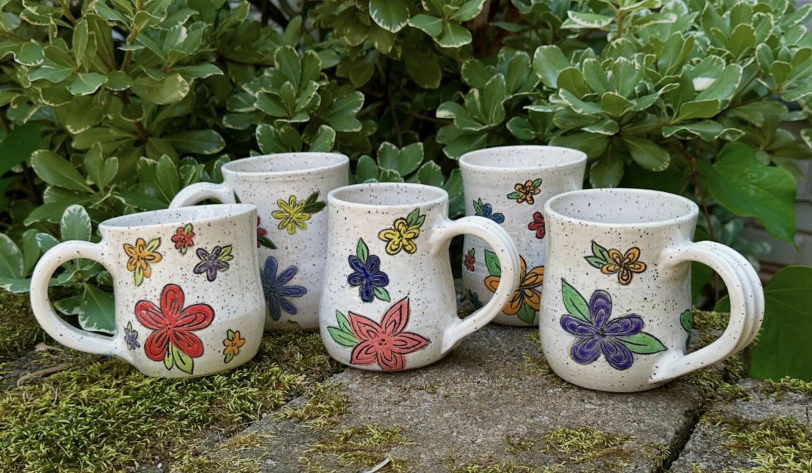Double Bloom Ceramics - Five ceramic mugs with colorful floral designs are arranged outdoors on a mossy stone surface with green foliage in the background.