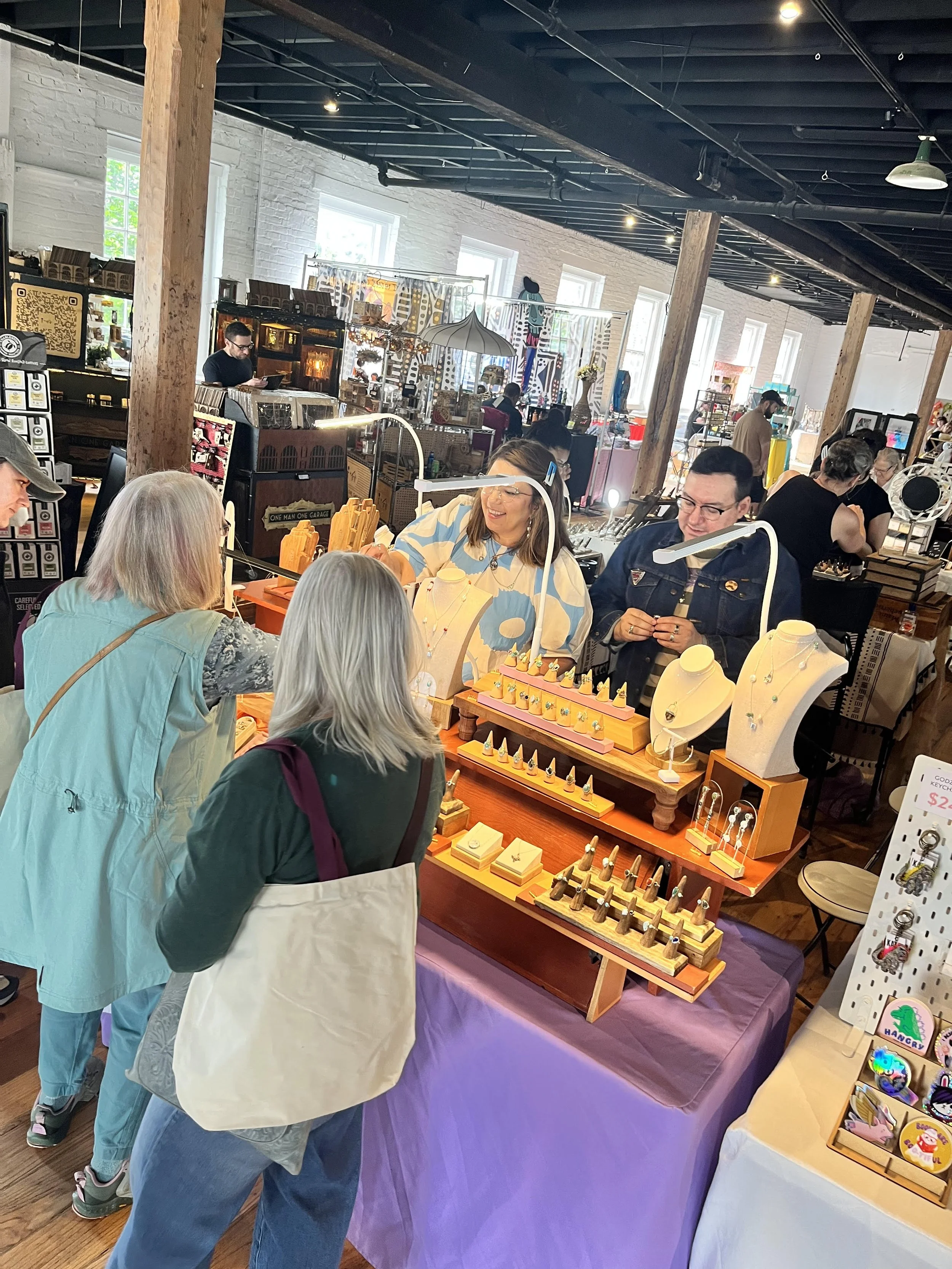 People browsing jewelry and accessories at an indoor market or craft fair with various stalls and displays.