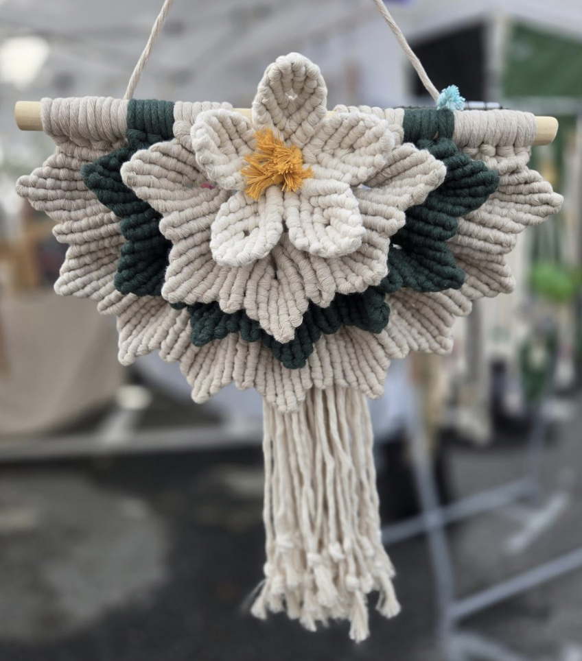 MacraYAY - Macramé wall hanging featuring a large cream-colored flower with yellow center, dark green leaves, and cream tassels hanging below.