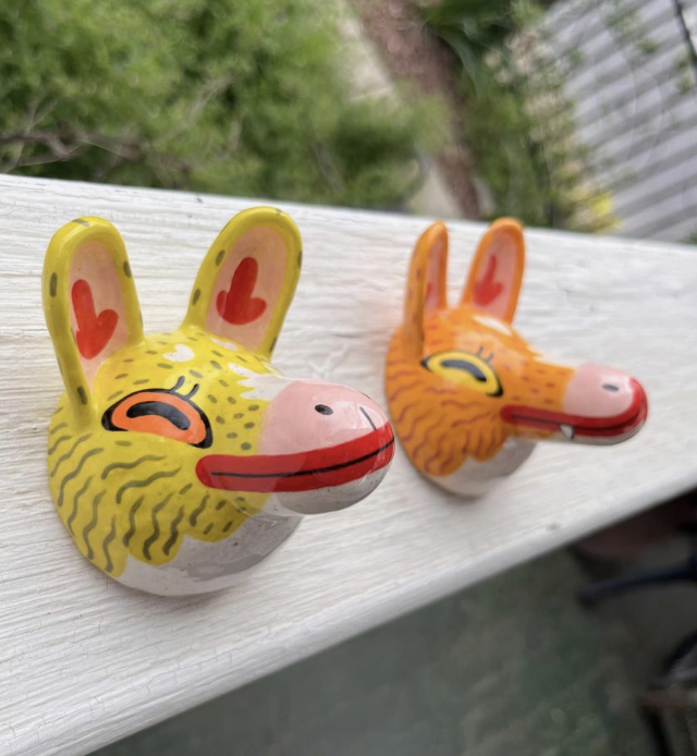 RJ Pesce - Two ceramic rabbit-shaped masks with colorful designs, one yellow with pink and orange accents and one orange with pink and yellow accents, are placed on a white wooden surface outdoors.