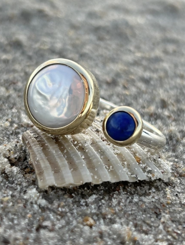 Heather Greenway Jewelry - Two rings with large gemstone settings, one with a pearlescent gemstone and the other with a dark blue gemstone, resting on a seashell on sandy ground.