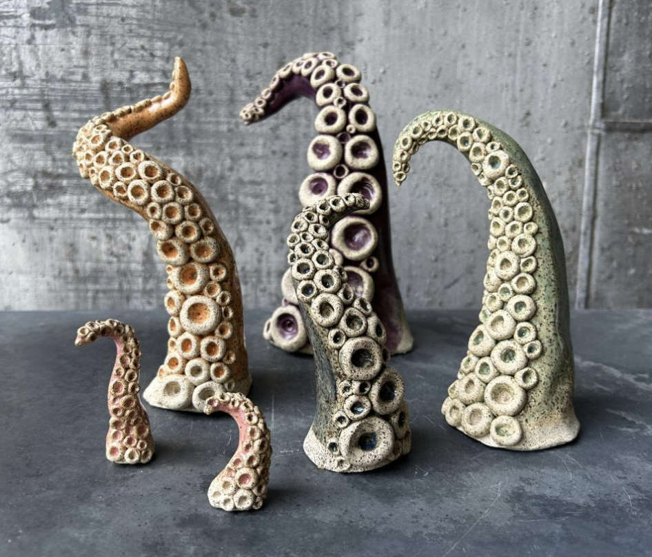 Hello Splendor - Ceramic octopus tentacle sculptures of various sizes with circular suction cups on a dark gray surface against a concrete wall.