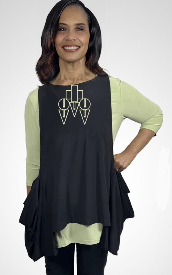 Lagenlooks - Smiling woman with dark hair in a middle part, wearing a lime green long-sleeve top and a black apron with geometric designs, standing with one hand on her hip against a plain light background.