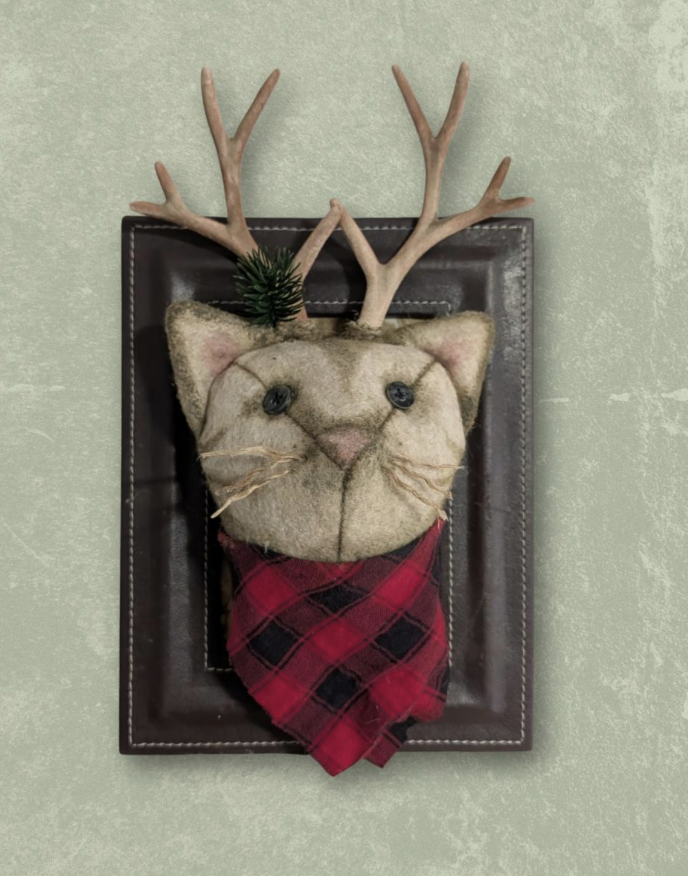 Birthcraft - Decorative cat face wall hanging with antlers, bouton eyes, pink nose, whiskers, wearing a red and black plaid bandana, mounted on a black frame on a light green wall.