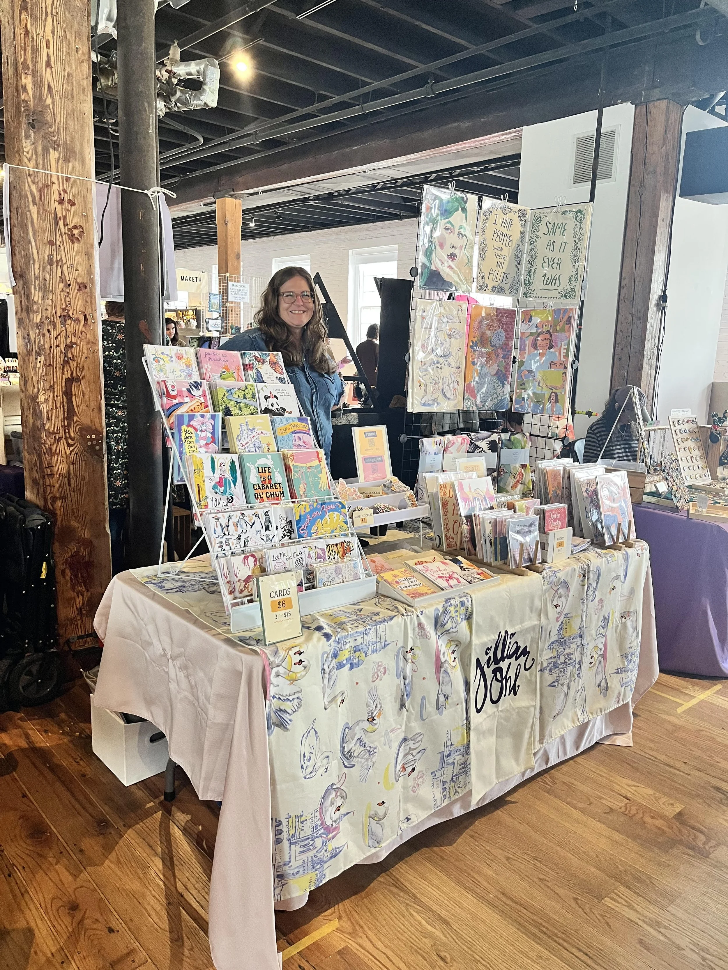A woman standing behind a table at an art and craft market booth, displaying various colorful greeting cards, prints, and artwork with handwritten price tags, in an indoor market space with wooden floors, exposed ceiling beams, and other stalls in the background.