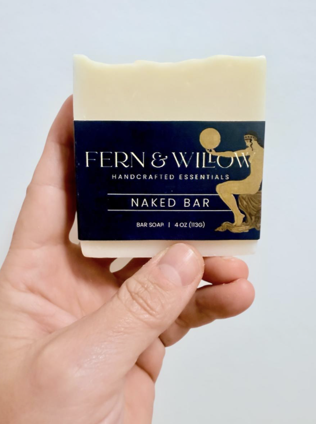 A hand holding a bar of soap with a navy blue label that reads 'Fern & Willow Handcrafted Essentials Naked Bar, Bar Soap, 4 oz, 113 g'. The label features an illustration of a mermaid holding a sphere.