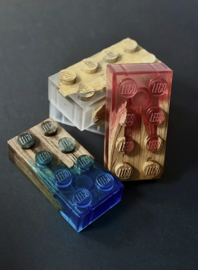 He's Here All Week 3D printing - Three LEGO brick displays with different colors and materials: a wooden base with blue translucent bricks, a marble-looking base with gray translucent bricks, and a wooden base with beige bricks, all set against a dar