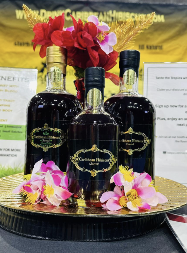 Three bottles of Caribbean Hibiscus syrup arranged on a gold platter, garnished with pink and yellow hibiscus flowers and golden ferns, with informational signs and a yellow banner in the background.