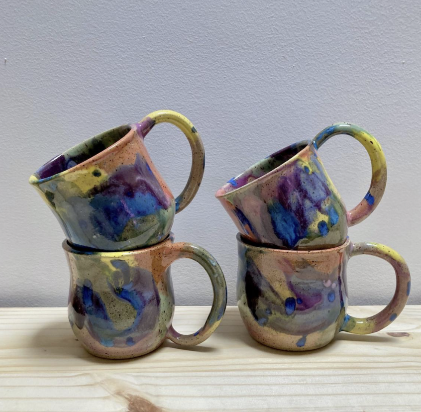 Katie Troisi Pottery - Four colorful ceramic mugs with abstract blue, yellow, pink, and green patterns, stacked in pairs on a light wooden surface against a plain wall.