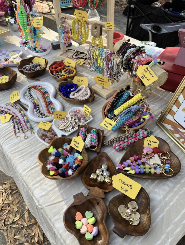 A table at an outdoor craft fair displaying colorful beaded jewelry, including bracelets, necklaces, earrings, and rings, arranged in wooden bowls and on a stand, with small yellow price tags.