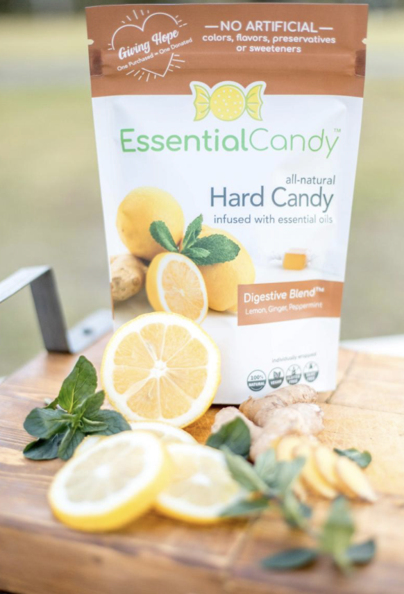 A package of Essential Candy all-natural hard candy infused with essential oils, featuring flavors like lemon, ginger, and peppermint, displayed with fresh lemon slices, ginger root, and mint leaves on a wooden surface.