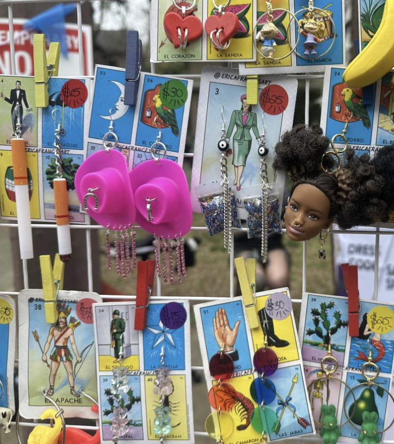 Erica Frances Art - A display of various earrings, keychains, and small accessories hanging on a grid at an outdoor market. The items include colorful earrings shaped like hearts, animals, and cartoon characters, along with keychains of animals and i