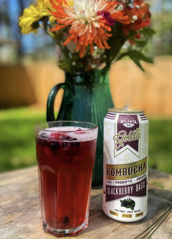 Cultured South Catering - A glass of dark purple berry kombucha next to a tall can of Blackberry Basil flavored kombucha on a wooden surface outdoors, with a green vase of orange, white, yellow, and purple flowers in the background.