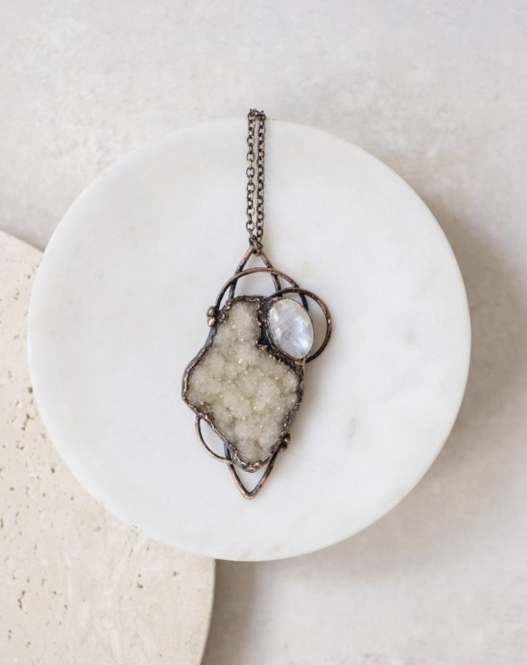 Pure Ritual - Asheville Jeweler - A pendant necklace with a wire-wrapped design, featuring a crystal geode slice and a small oval-shaped iridescent stone, resting on a white round surface.