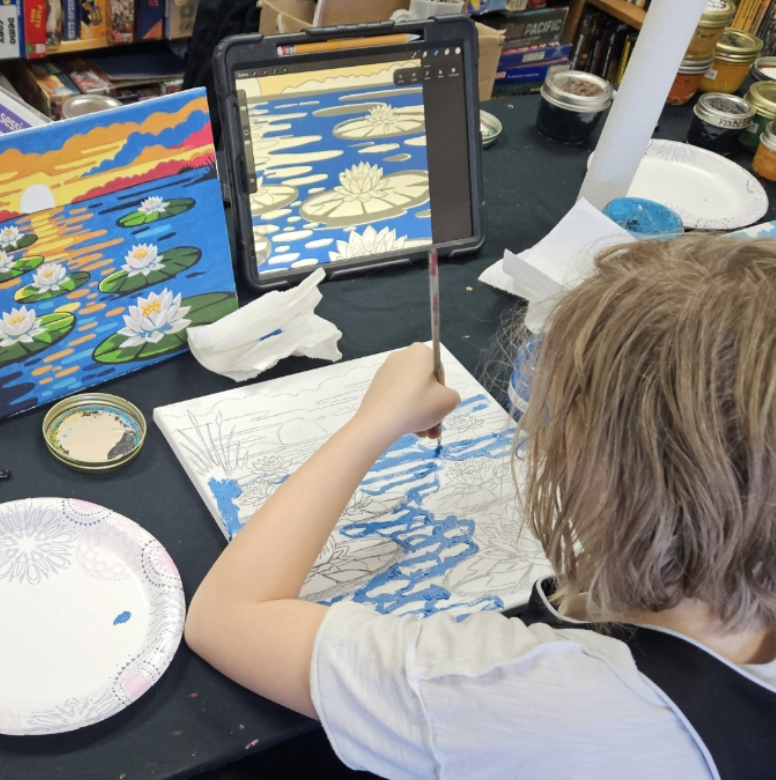 Child painting a water lily scene inspired by a digital reference on a tablet, with finished painting of water lilies and a sunset scenery on display.