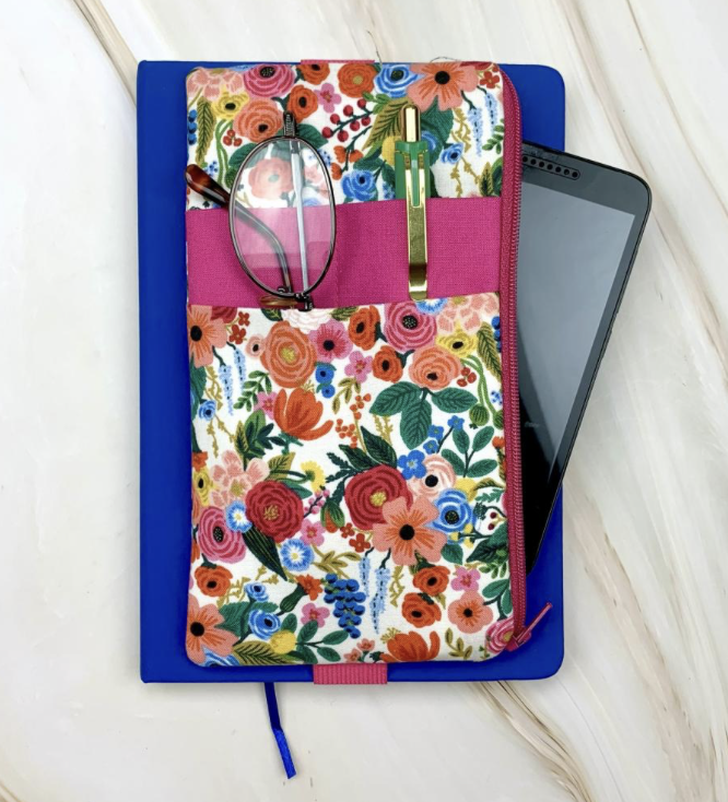 Joy Town Studio - A tablet partially inside a floral fabric case, resting on top of a blue notebook on a white marble surface. The case has a pink elastic band and includes a pair of glasses and a yellow pen.