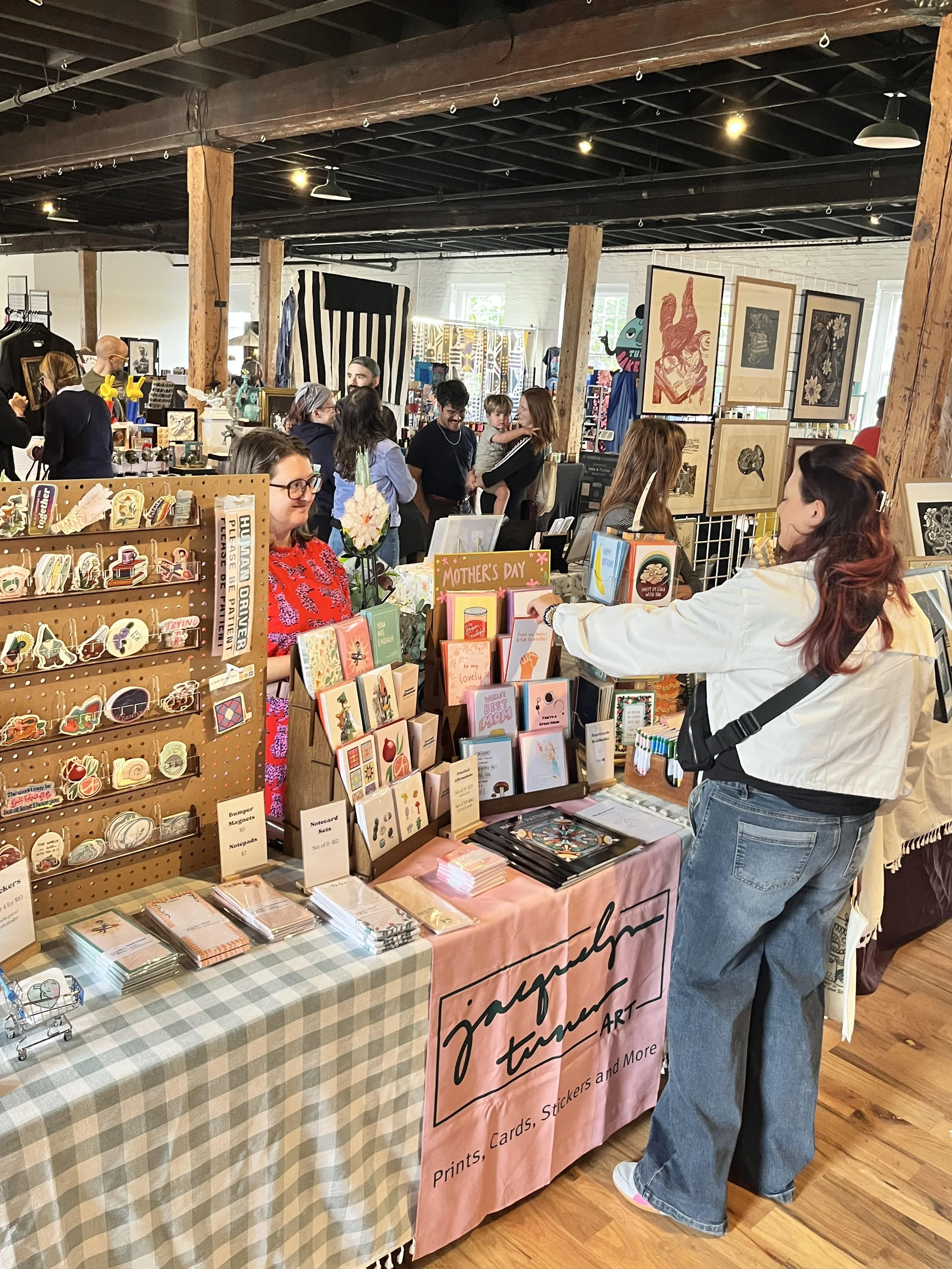 A bustling indoor market with various booths. In the foreground, a woman in a white jacket is browsing items on a table that sells cards, stickers, and prints. A woman with glasses in a red dress stands behind the table. In the background, other customers and vendors browse and interact, with artwork displayed on boards and walls, and wooden beams overhead.