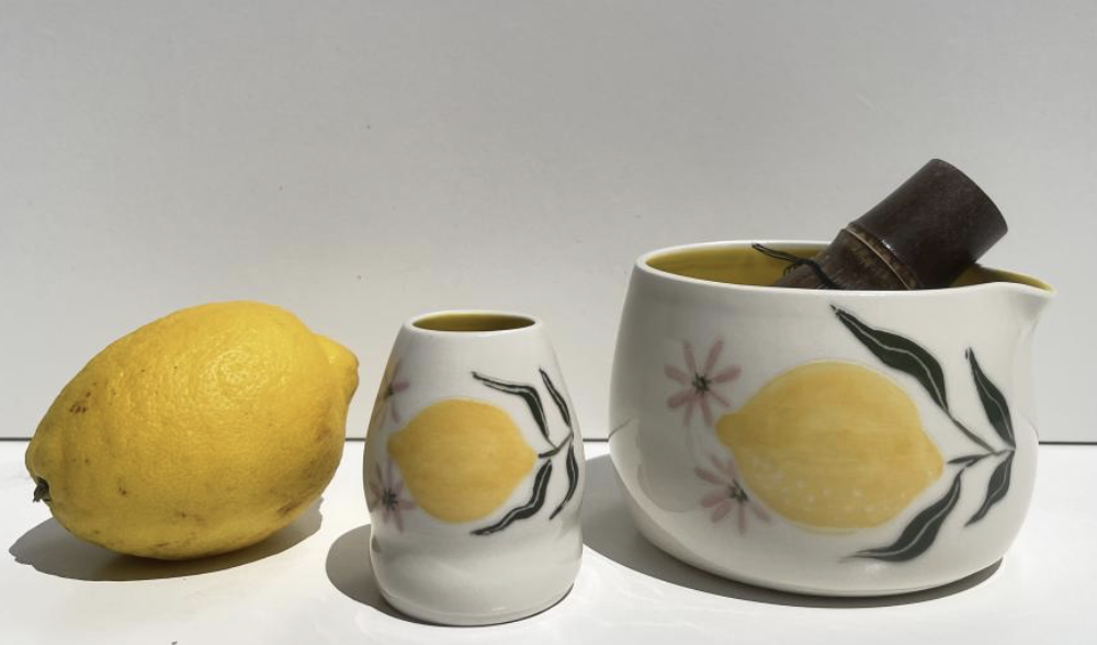 LB Ceramics - A bright yellow lemon, a small ceramic cup with lemon and floral design, and a larger ceramic pitcher with a similar lemon and floral design containing a hand-rolled copper object with a black handle.
