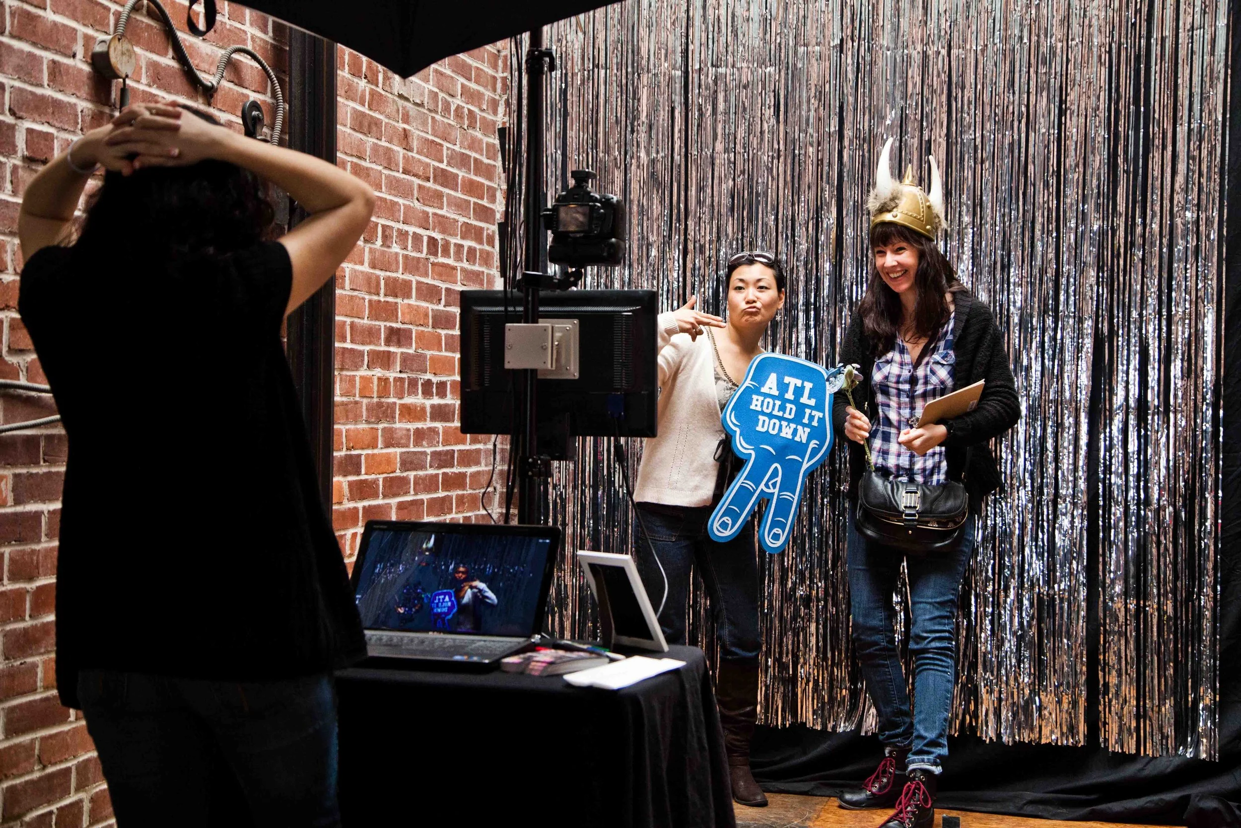 Two women are standing in front of a shiny, black, tinsel backdrop at a photo booth. One woman is holding a blue sign that says 'ATL HOLD IT DOWN' and a single flower, while the other woman is wearing a Viking helmet with horns and smiling. A person is taking their picture, with a camera and a laptop visible in the foreground.